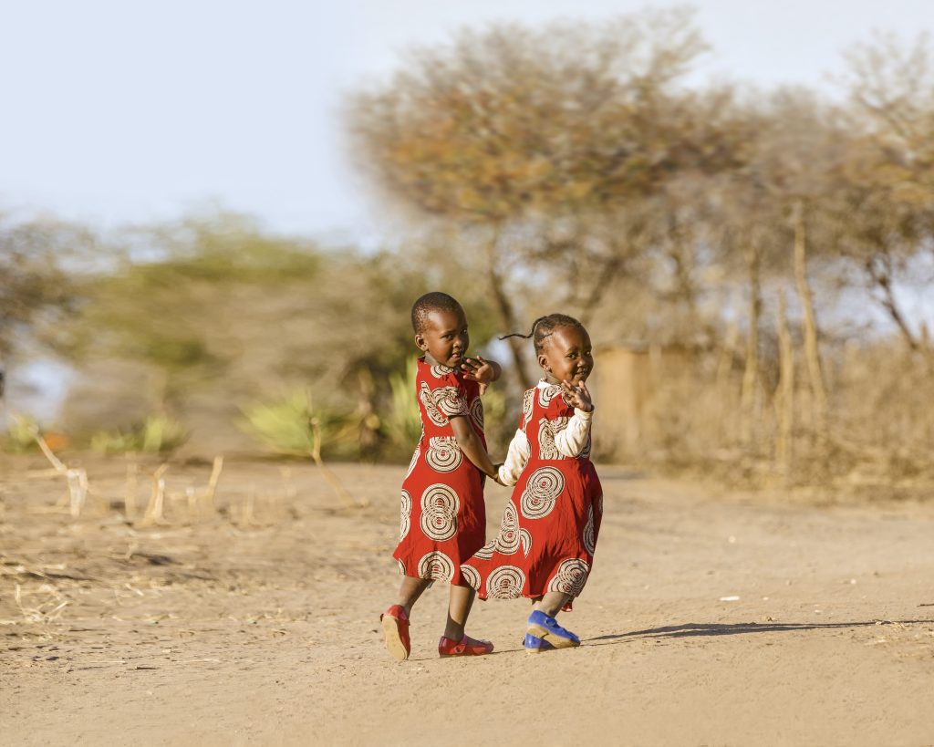 Massai Mädchen in rot gemusterten Kleidern lächeln und winken in die Kamera