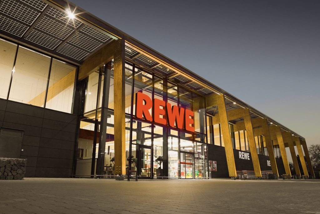 Bild der Fassade eines REWE Supermarktes in der Dämmerung, das Gebäude ist angestrahlt