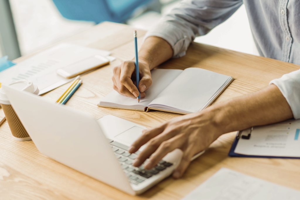 Einen Plan machen. Beschäftigter Mann stützt sich mit den Ellbogen auf den Arbeitsplatz und arbeitet mit dem Computer, schreibt Informationen in ein Notizbuch