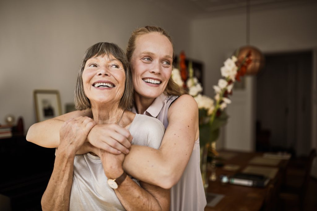 Fröhliche reife Frau umarmt ältere Mutter zu Hause und schaut weg. Porträt einer älteren Mutter und einer Tochter mittleren Alters, die zusammen lächeln.