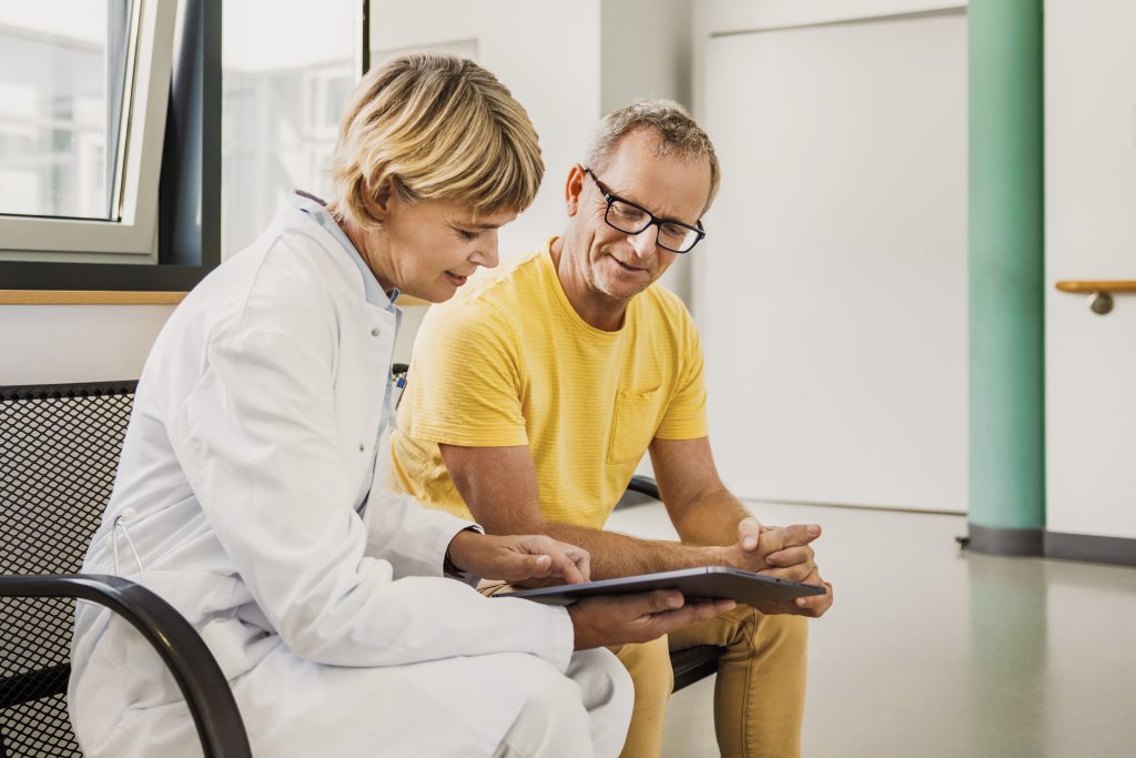 Eine reife Ärztin und ein älterer Patient sitzen in einem Krankenhaus zusammen und sehen sich Informationen auf einem digitalen Tablet an. Beide sind in ein nachdenkliches Gespräch vertieft, das auf einen kooperativen Ansatz bei der Patientenversorgung hindeutet.