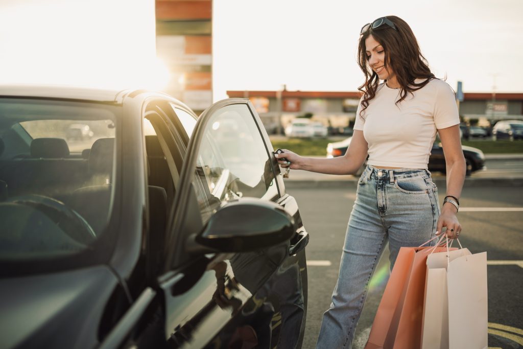 Lächelnde trendige Mädchen stehen auf Parkplatz mit Einkaufstaschen in den Händen und in ihr Auto. Porträt einer glücklichen modischen jungen Frau, die ihr Auto nach einem Einkaufsbummel im Einkaufszentrum betritt.