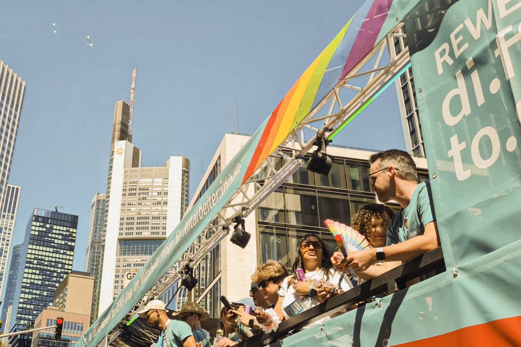 Menschen auf einem Wagen mit Regenbogenbanner und REWE Di.to Aufschrift vor der Skyline Frankfurts
